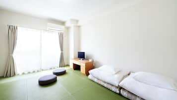 A minimalist hotel room with a green tatami mat floor, a bed with white bedding, a small wooden desk with a television, and a window with curtains.