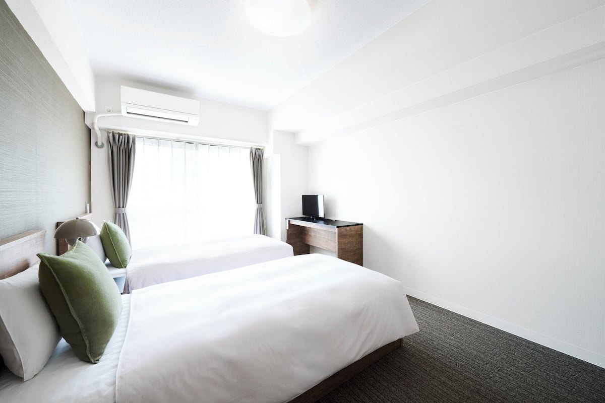 A hotel room with two beds, a desk, an air conditioning unit, and a window with curtains.