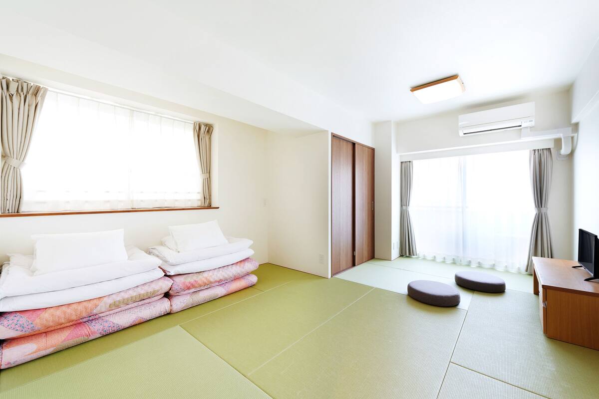 A room with tatami mats, a bed with white bedding, a desk with a monitor, and an air conditioning unit.