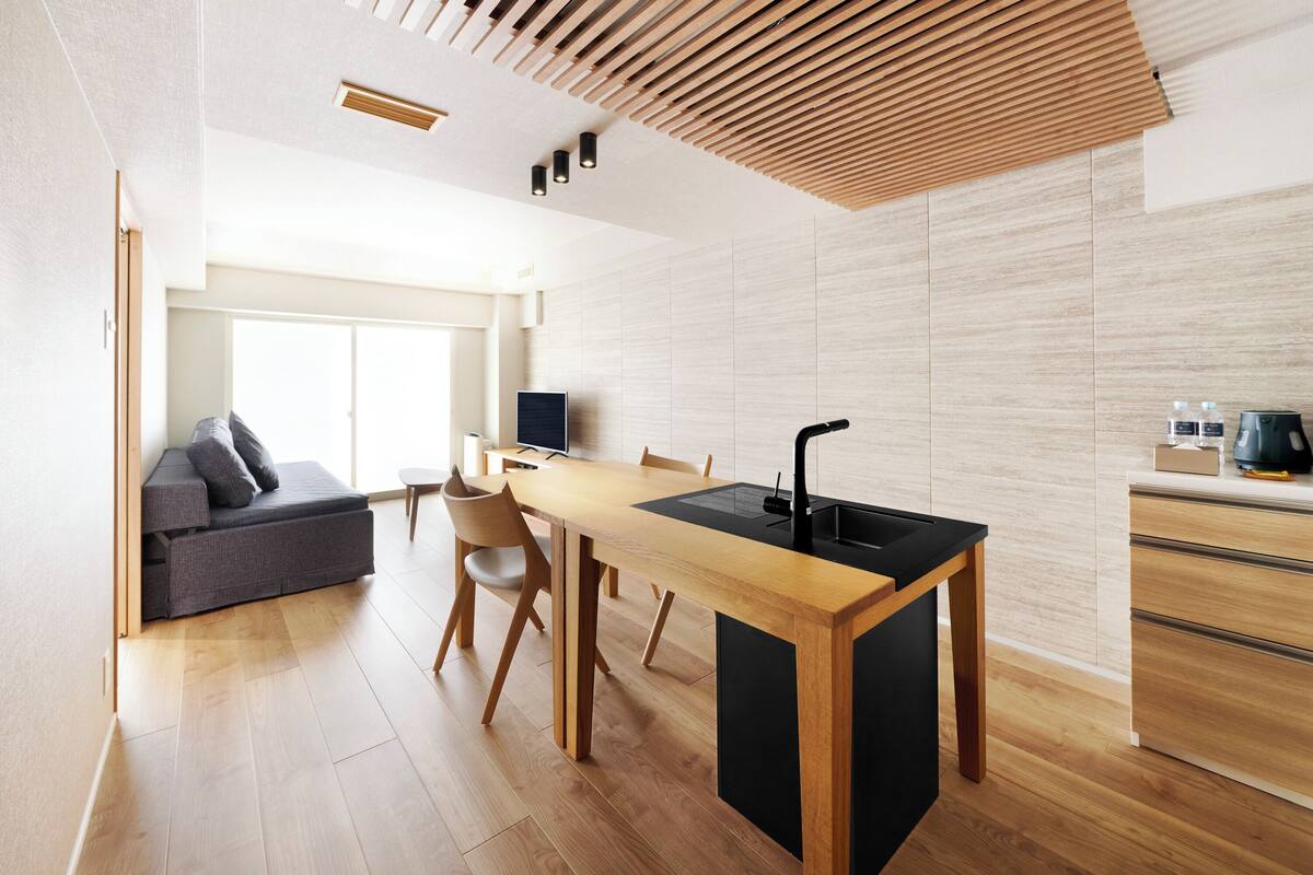 A modern kitchen with a wooden table and black countertop, a grey sofa, and a TV in the background.