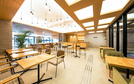 A modern dining area with wooden tables and chairs, a bar area, and large windows allowing natural light.
