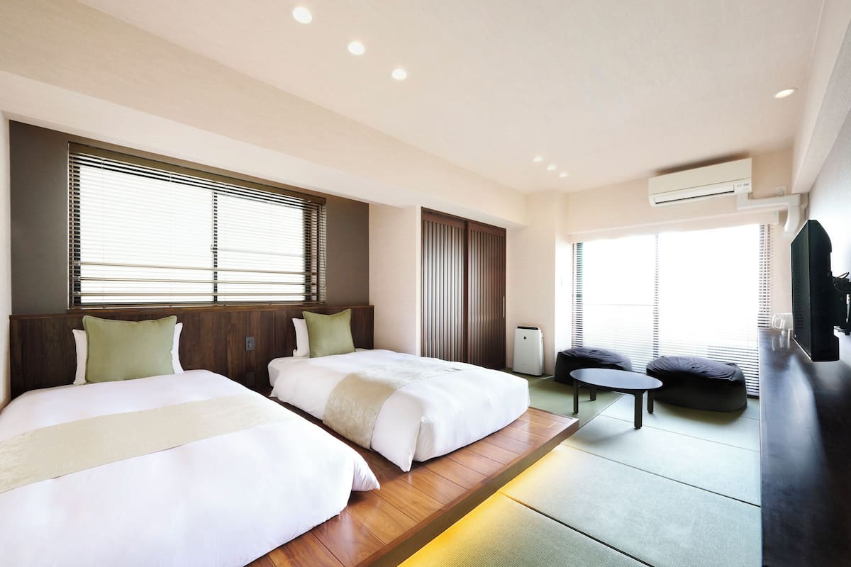 A hotel room with two beds, a small sofa, a TV, and a ceiling air conditioner.