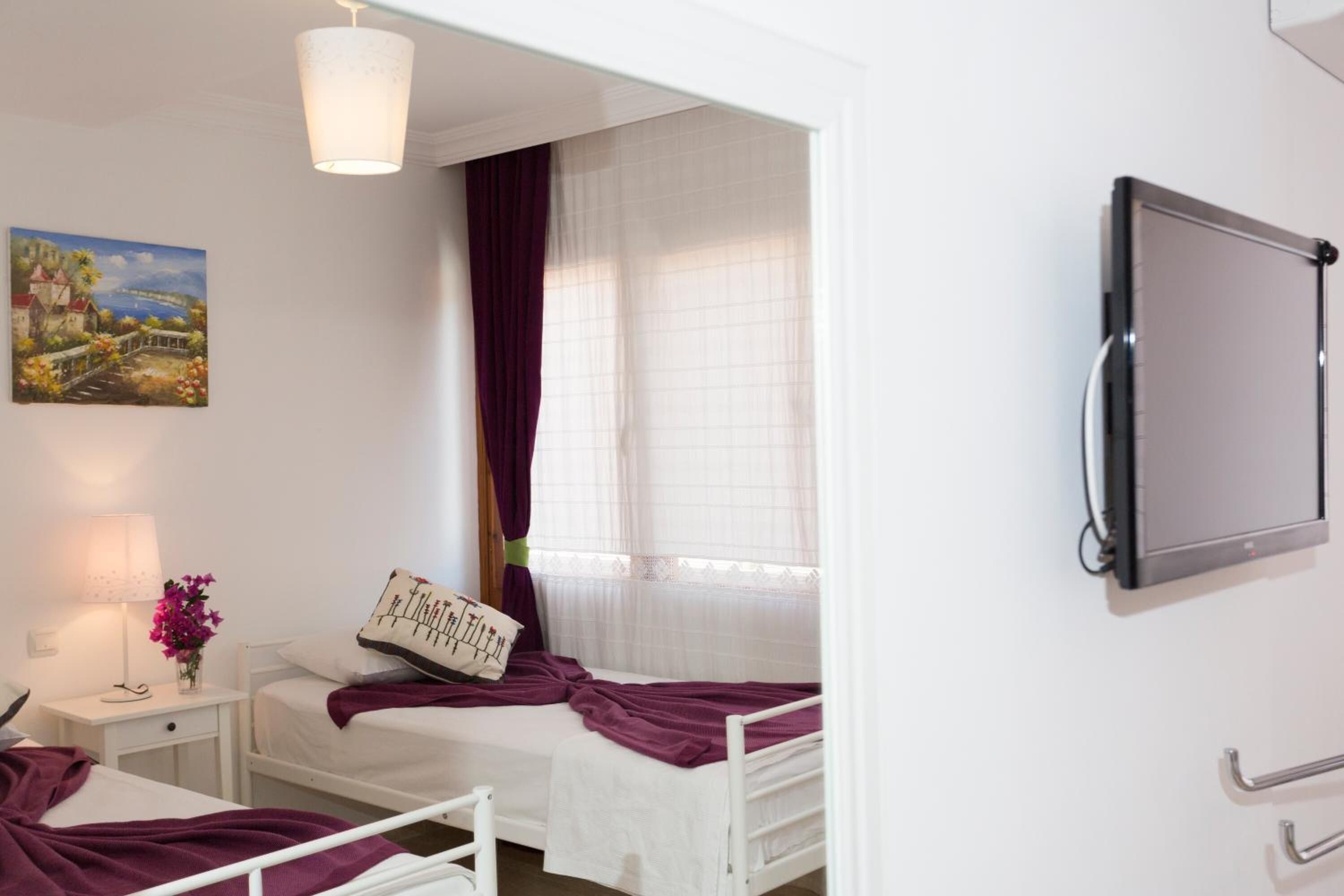 A bedroom with two beds, a bedside table with a lamp, a painting on the wall, and a window with curtains.