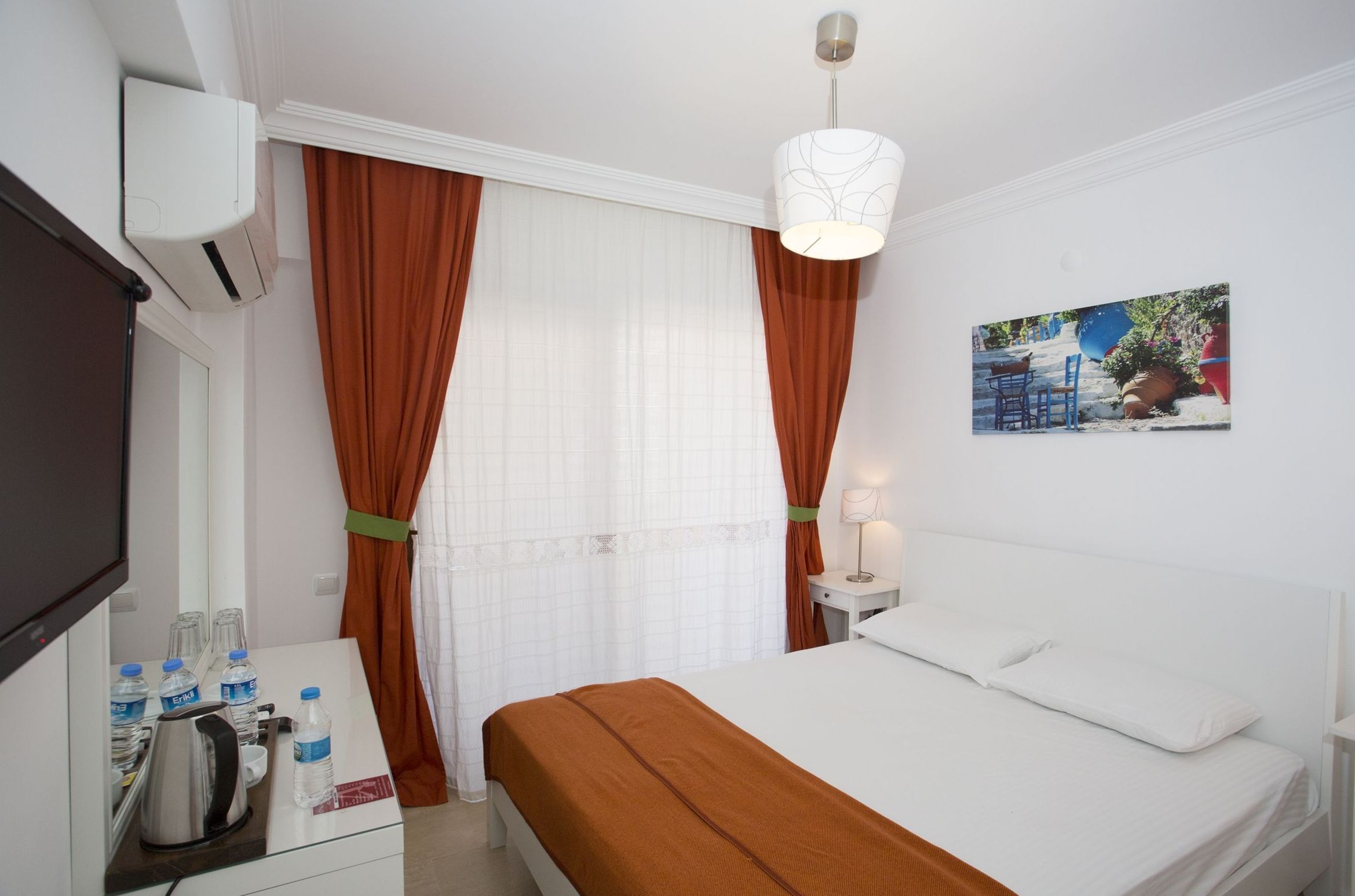 A hotel room with a bed, a desk with a kettle and water bottles, a TV, and a window with curtains.