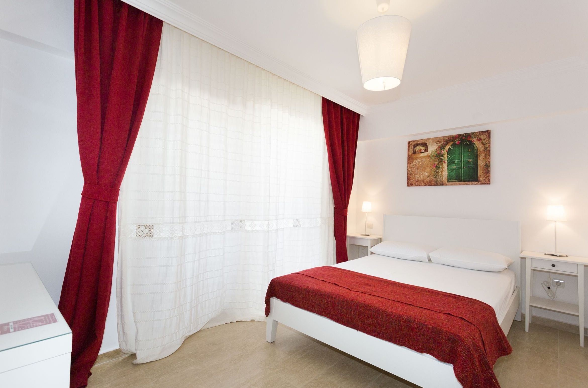 A neatly arranged bedroom with a bed, white curtains, a red blanket, a wall painting, and bedside lamps.