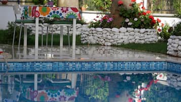 Zona de piscina con borde de azulejos, una banca con mantel de flores coloridas y una pared de piedra con flores.
