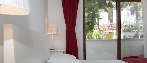 Habitación de hotel con cama, lámpara, ventana con vistas a follaje y balcón.