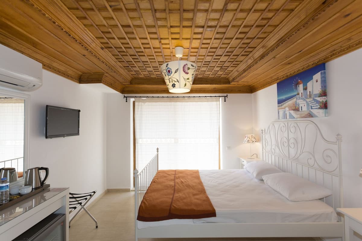 A bedroom with a wooden ceiling, a bed with white bedding, a painting on the wall, and a window with blinds.