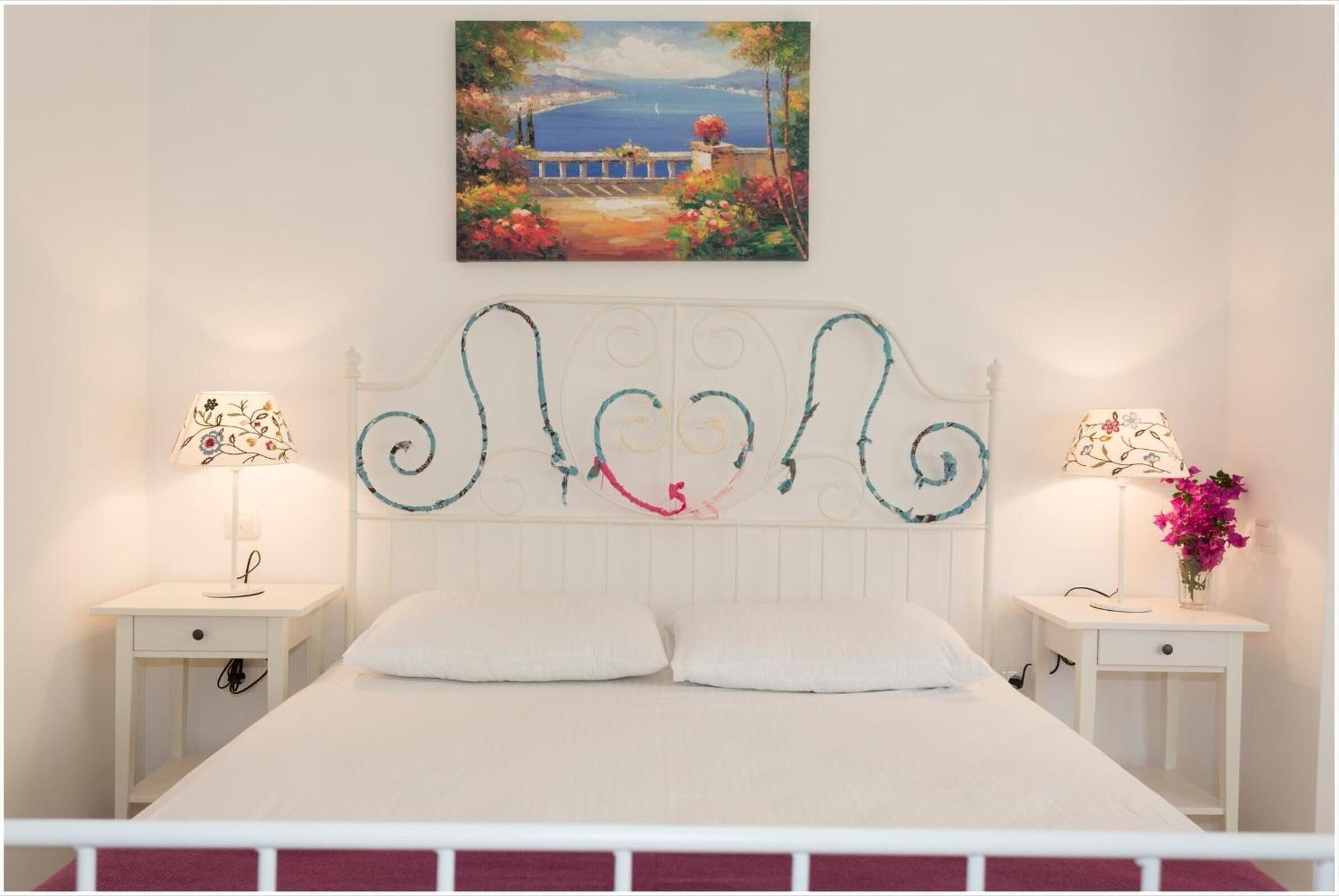 Un dormitorio con una cama blanca, dos mesitas de noche, un cabecero decorativo y un cuadro de una escena costera.