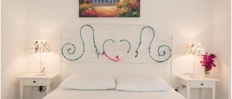 Un dormitorio con una cama blanca, dos mesitas de noche, un cabecero decorativo y un cuadro de una escena costera.