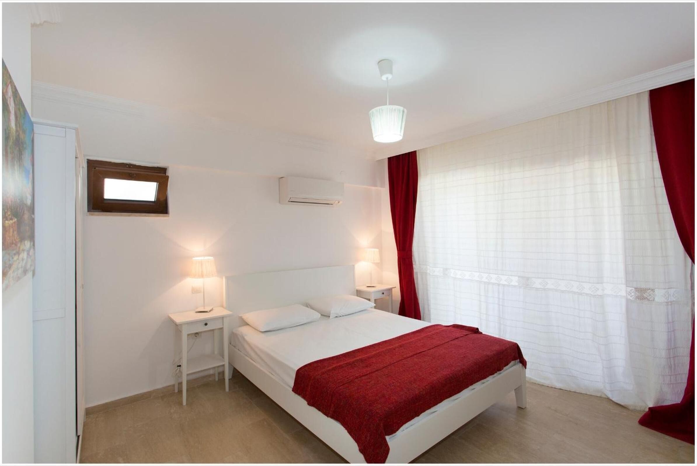 A bedroom with a bed, bedside lamps, a window with curtains, an air conditioning unit, and a ceiling light fixture.