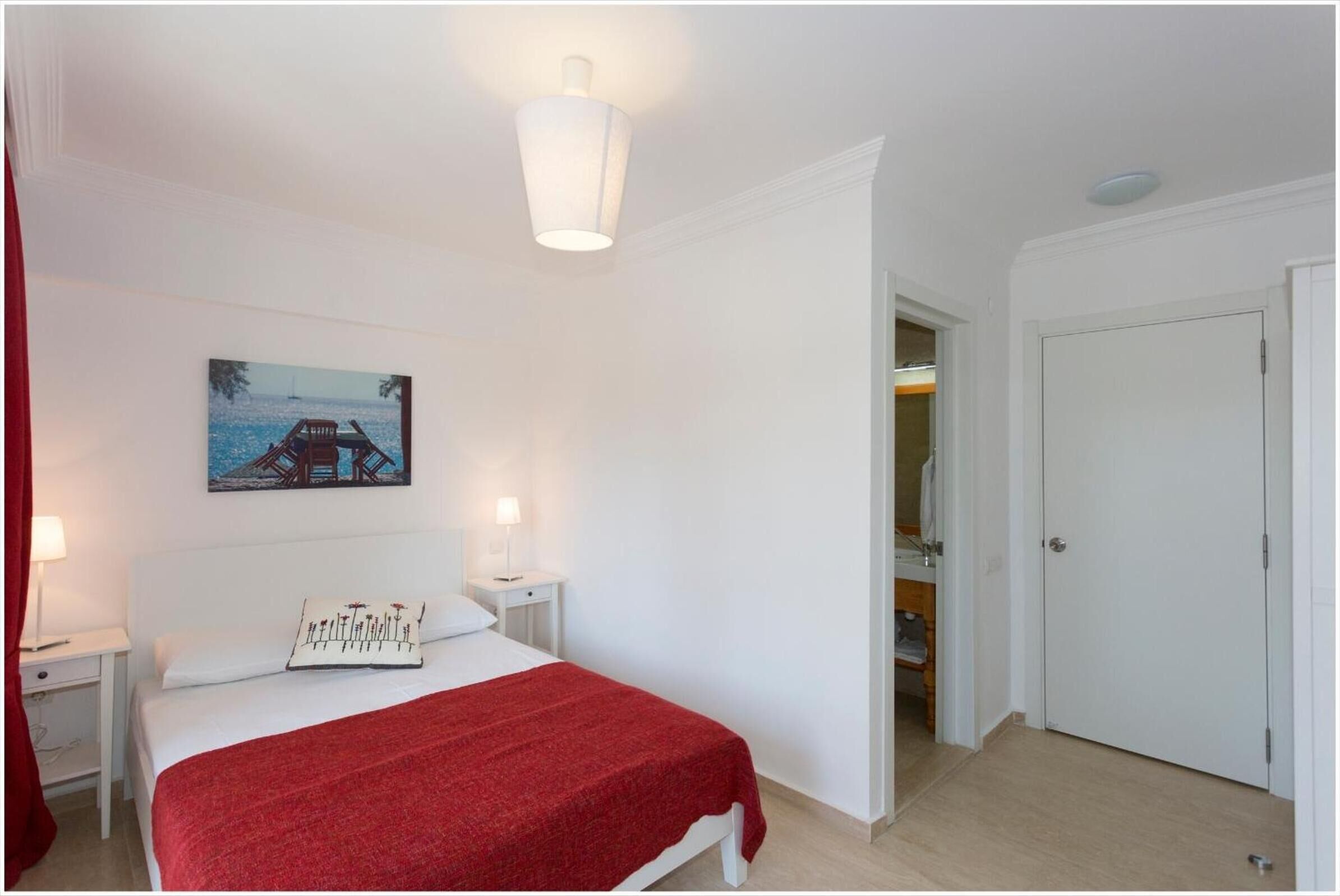 Un dormitorio con cama, mesita de noche, lámpara, un cuadro y una puerta.
