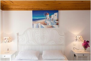 Un dormitorio con una cama blanca, dos mesitas de noche con lámparas, un cuadro que muestra una escena costera y un techo de madera.