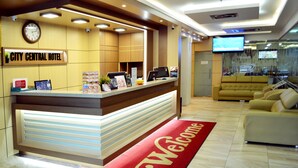 Zona de recepción de hotel con mostrador, reloj, televisor y un felpudo rojo.