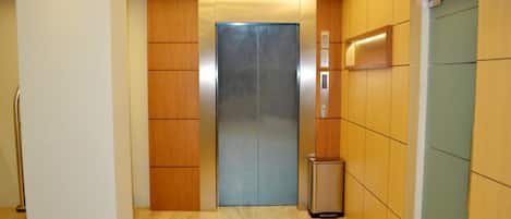 A modern elevator with a metallic door, wooden paneling on the walls, and a trash bin to the right.