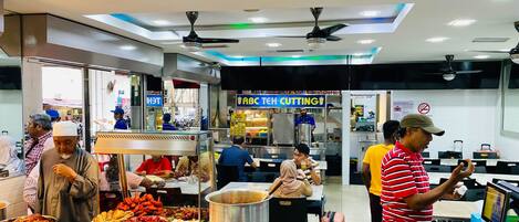 A food court with various food stalls, a ceiling fan, and a digital menu display.