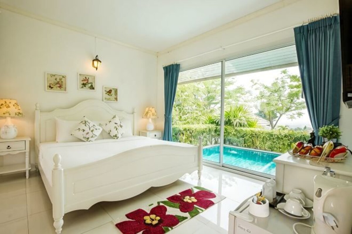 Un dormitorio con cama, mesitas de noche, una ventana con cortinas y vistas a una piscina, además de una pequeña cocina.