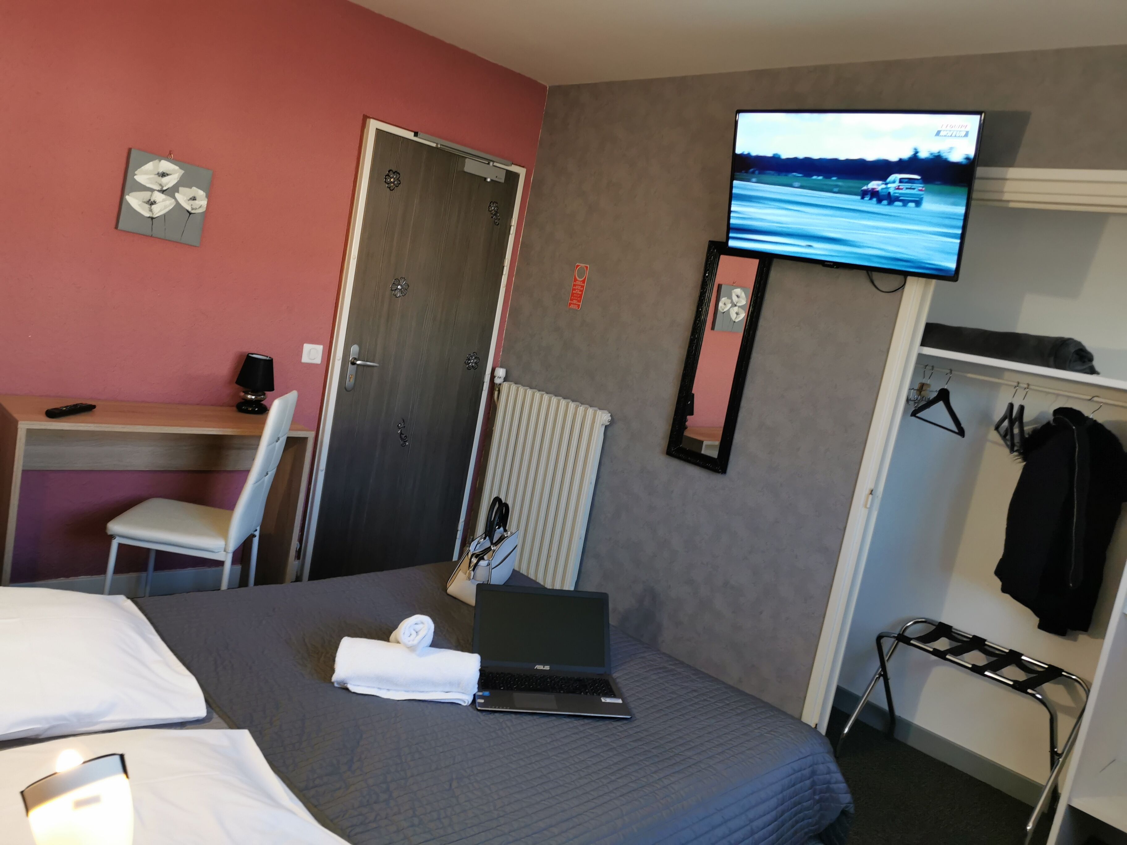 A hotel room with a bed, a desk with a chair, a laptop, a TV on the wall, and a coat rack.