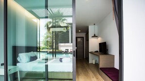 A modern hotel room with a glass wall, a bed, a small desk, a TV, and a wooden floor. - Chaweng Villawee (Koh Samui)