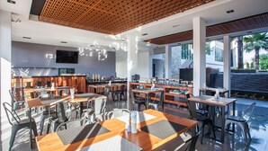 A modern dining area with multiple tables and chairs, a kitchen in the background, and large windows offering views of the outdoors. - Chaweng Villawee (Koh Samui)