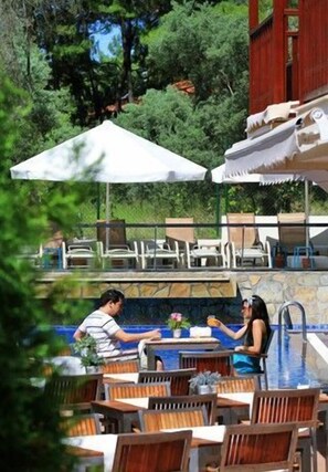 Zona de piscina al aire libre con sillas y mesas de madera, rodeada de vegetación y un edificio rojo.