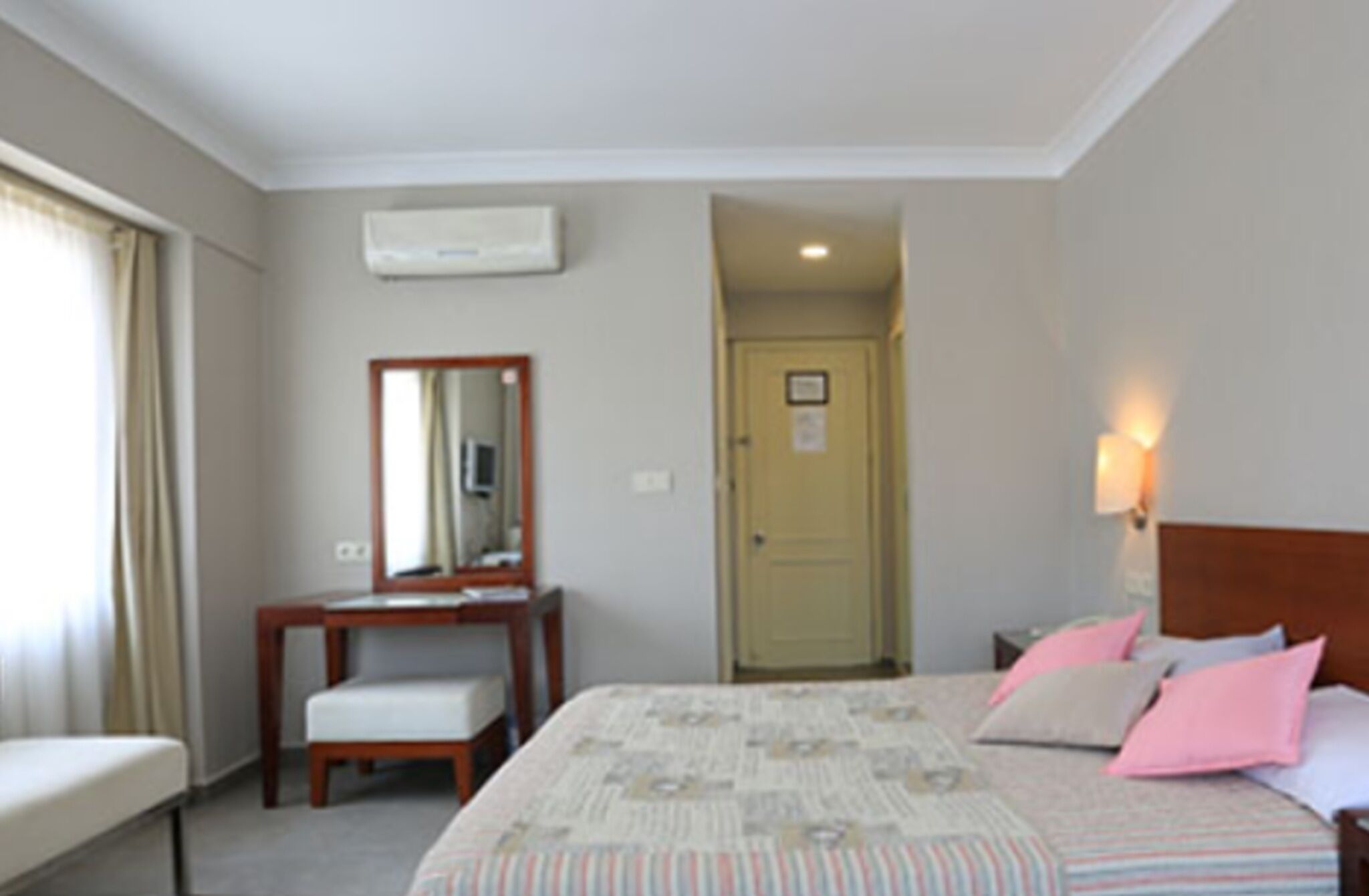 A hotel room with a bed, a desk, a chair, a mirror, a door, and a wall-mounted air conditioner.