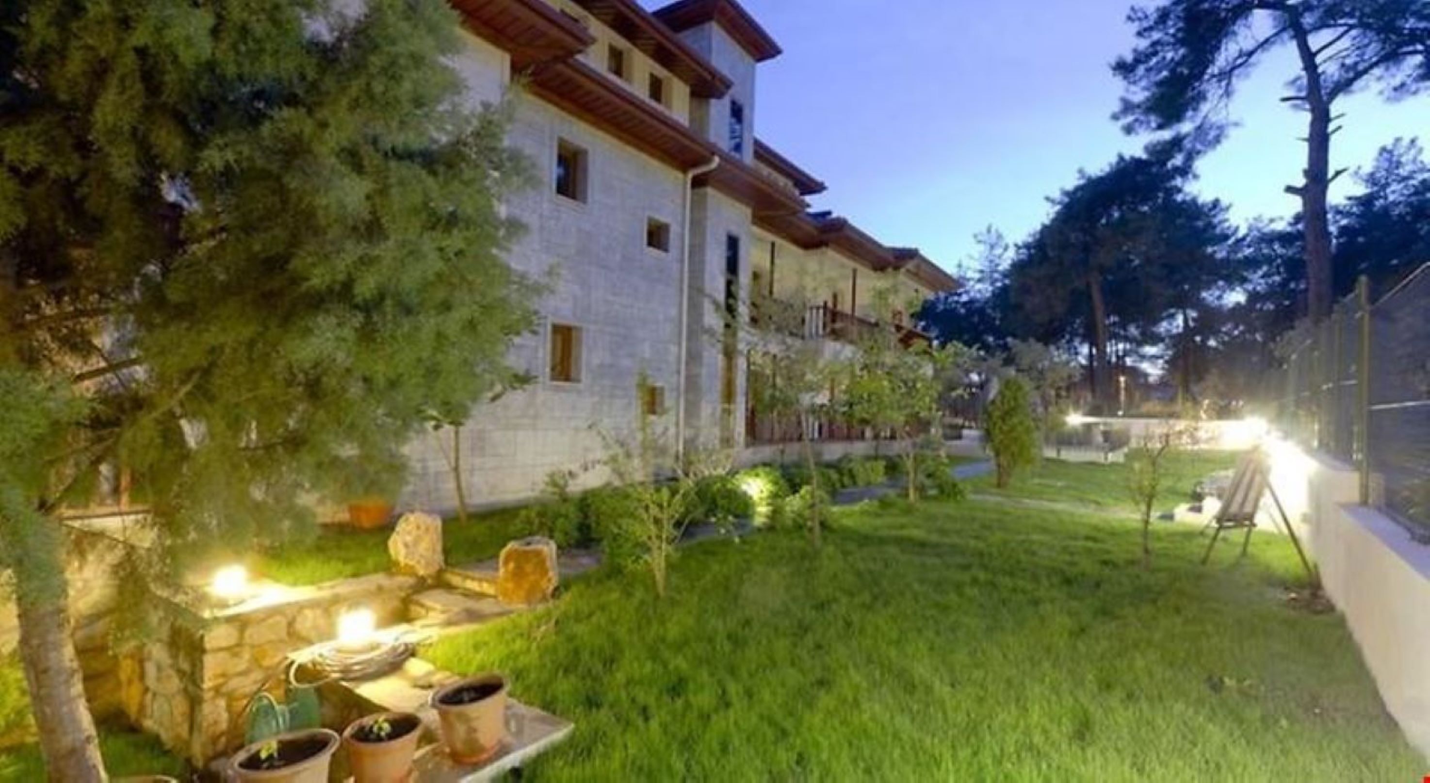 A two-story building with a stone facade and a red-tiled roof. There is a well-maintained lawn in front of the building, and a small garden area with a stone bench and potted plants.