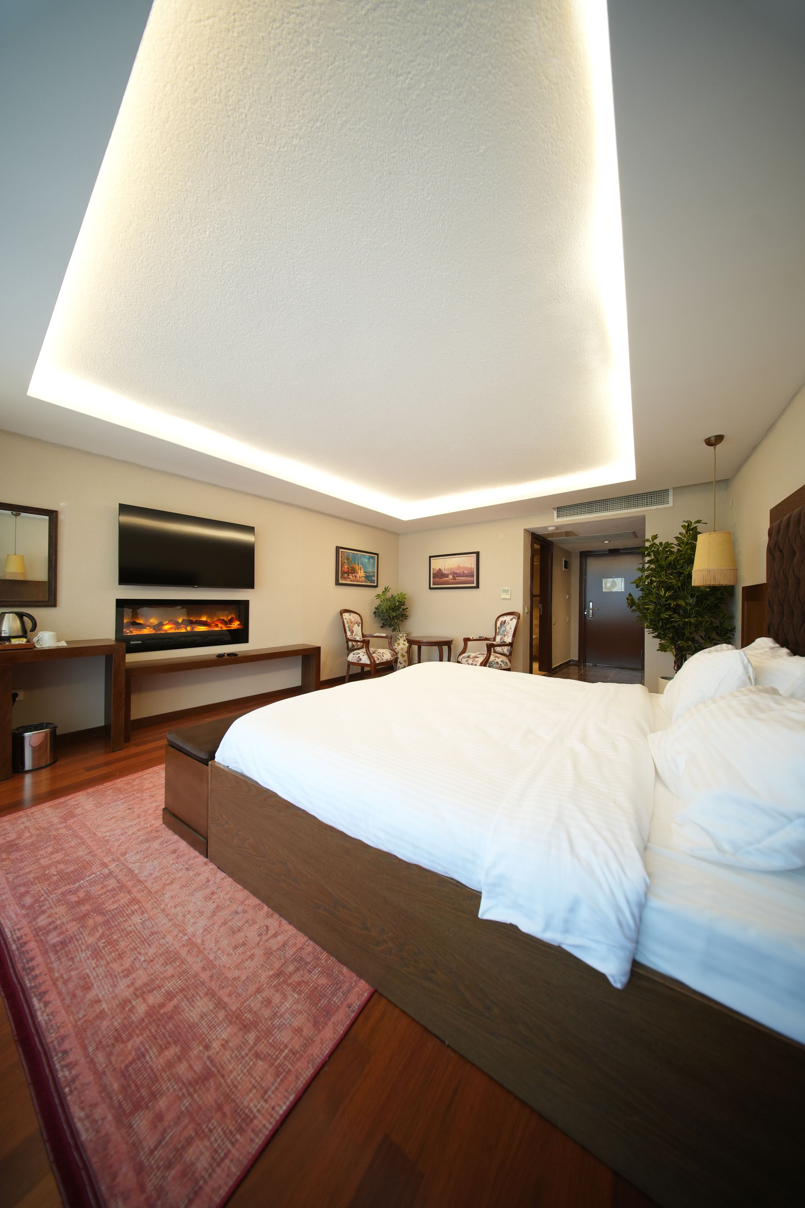 A modern hotel room with a large bed, a flat-screen TV, a fireplace, and a ceiling light fixture.
