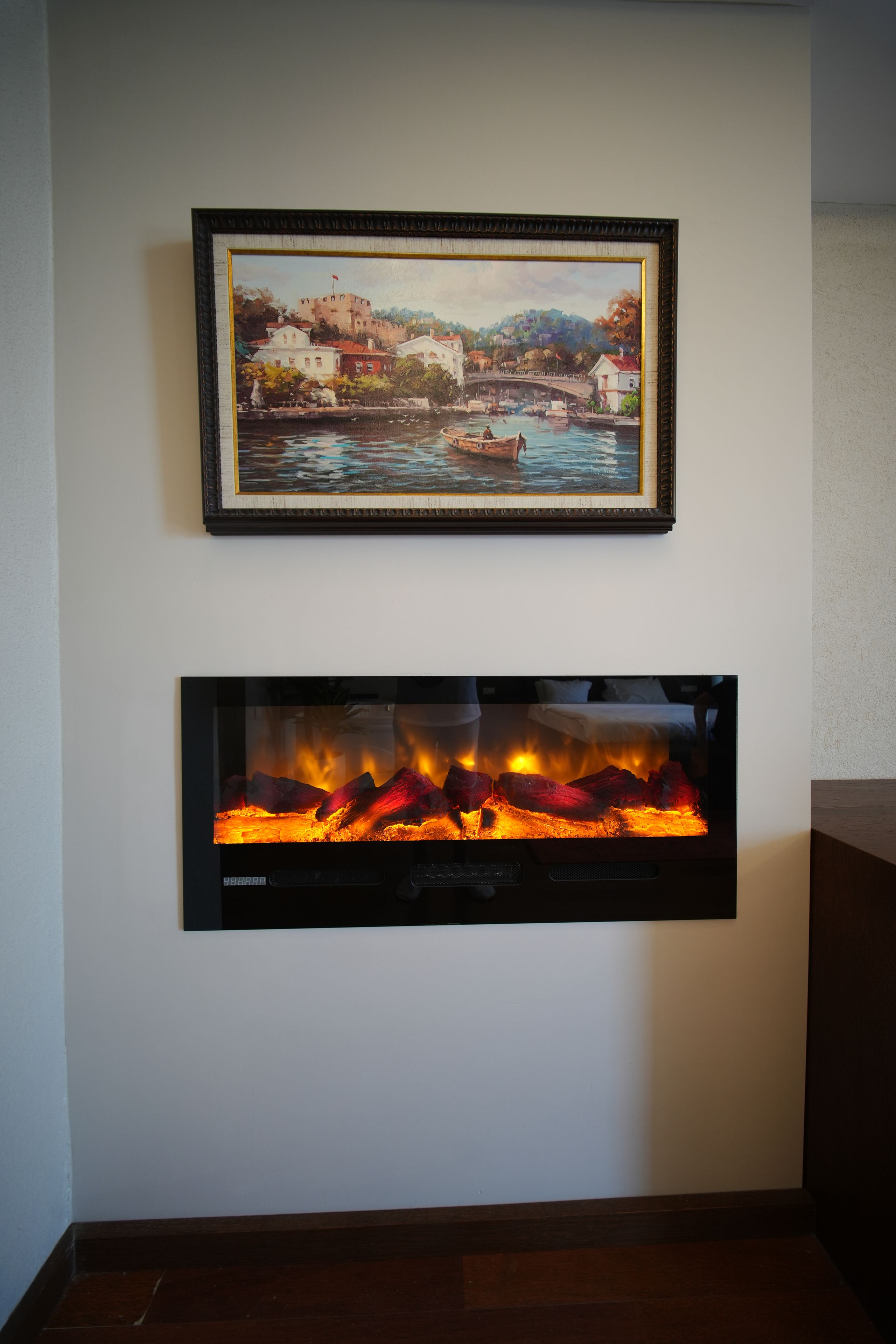 A framed painting of a coastal town and a modern fireplace below it.