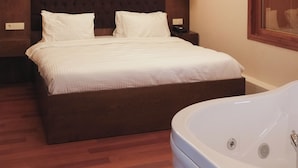 A hotel room with a large bed, a wooden headboard, two bedside lamps, a large window with blinds, and a white bathtub.