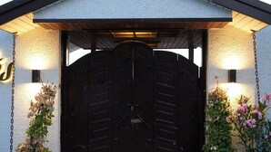 A building with a dark double door, a white exterior, and a roof with a gable. There are two potted plants, one on each side of the door.