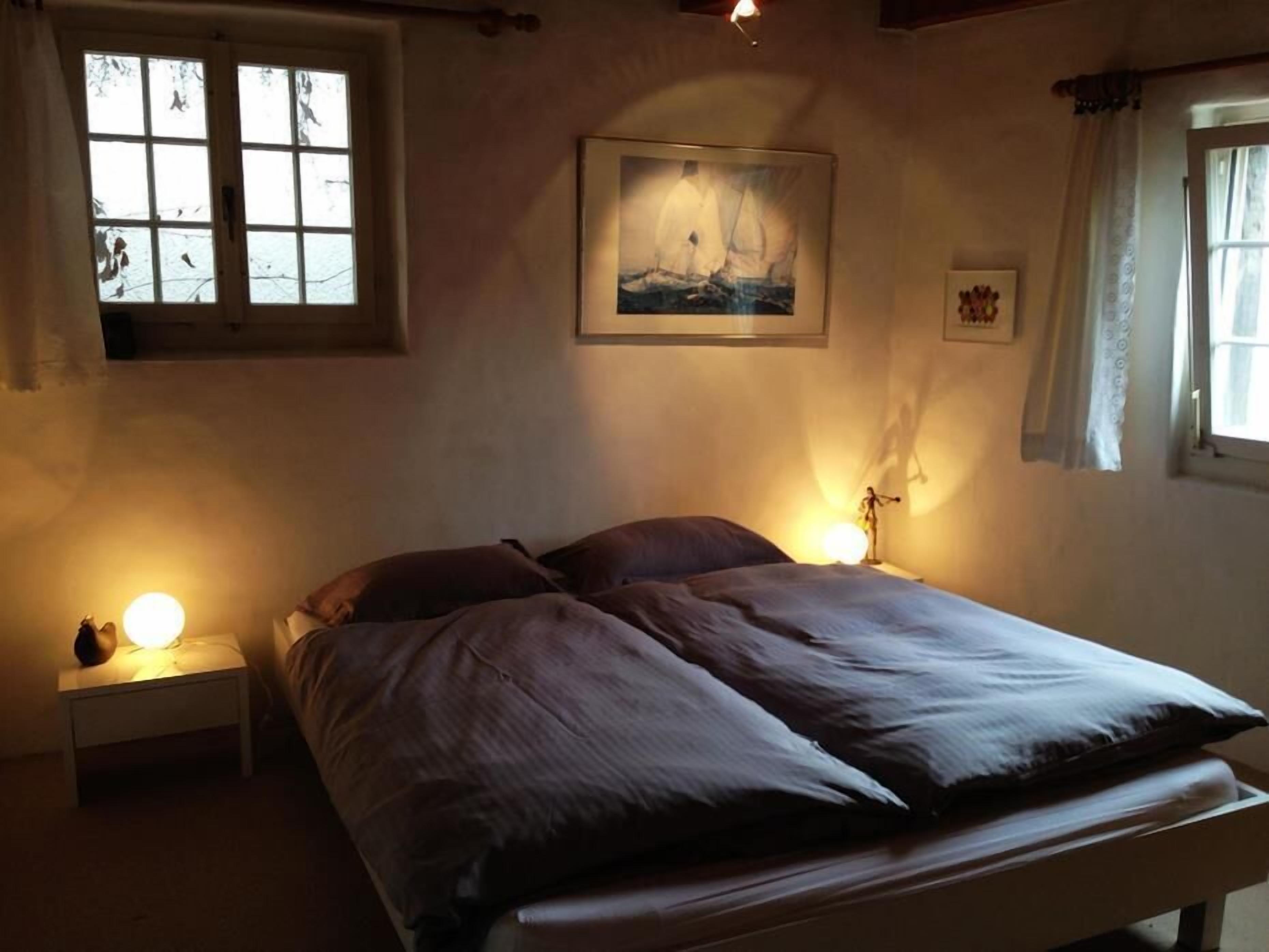 Un dormitorio con una cama, dos mesitas de noche, un cuadro colgado en la pared y una ventana con cortinas.