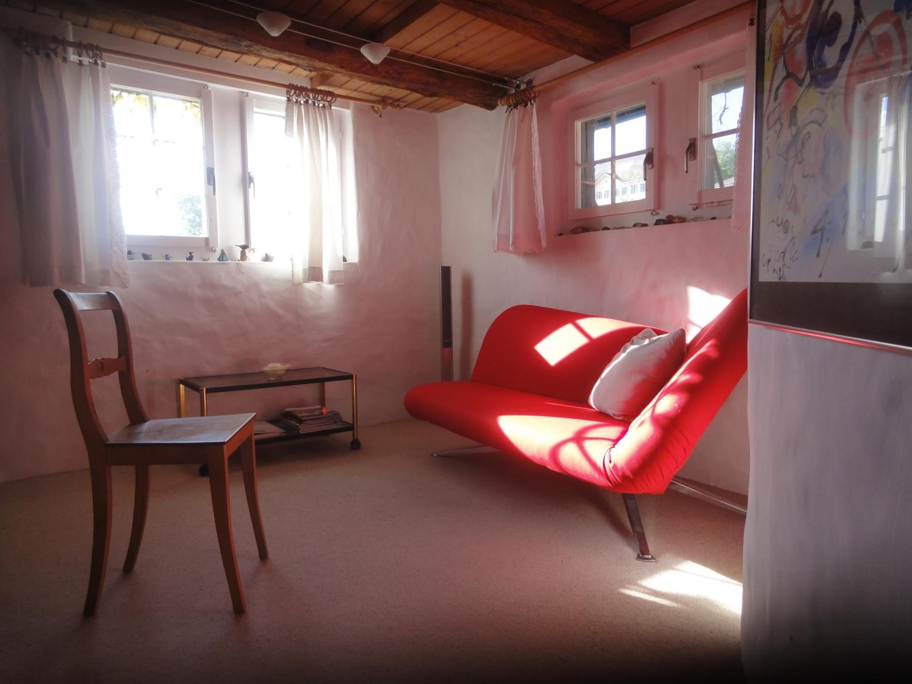Una habitación con techo de madera, un sofá rojo, una silla de madera, una mesita pequeña y un refrigerador.