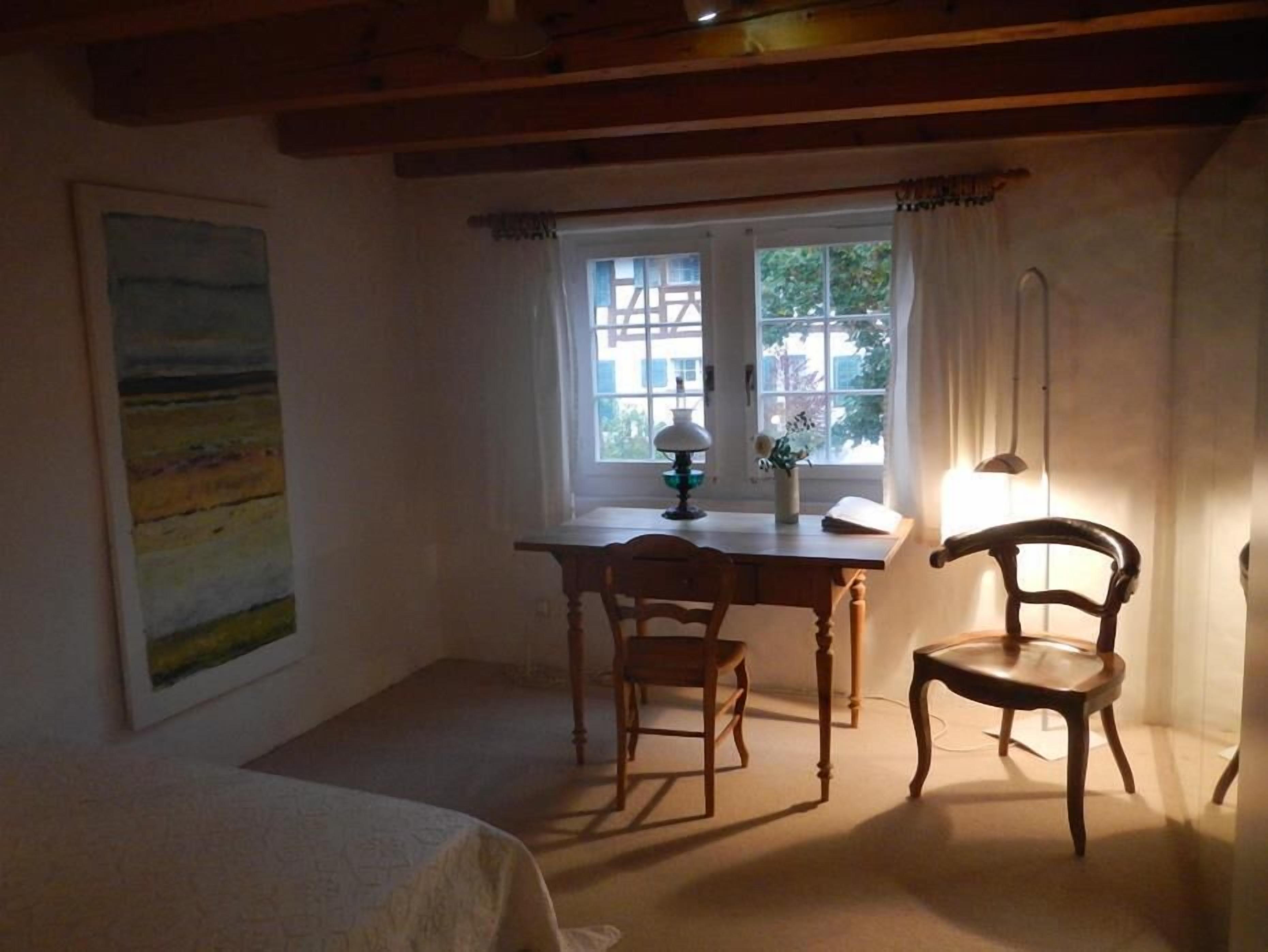 A cozy room with a bed, a desk with a chair, a window with curtains, and a painting on the wall.