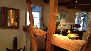 Un interior rústico con vigas de madera a la vista, una silla de madera, una mesa con una vela y un cuadro en la pared.
