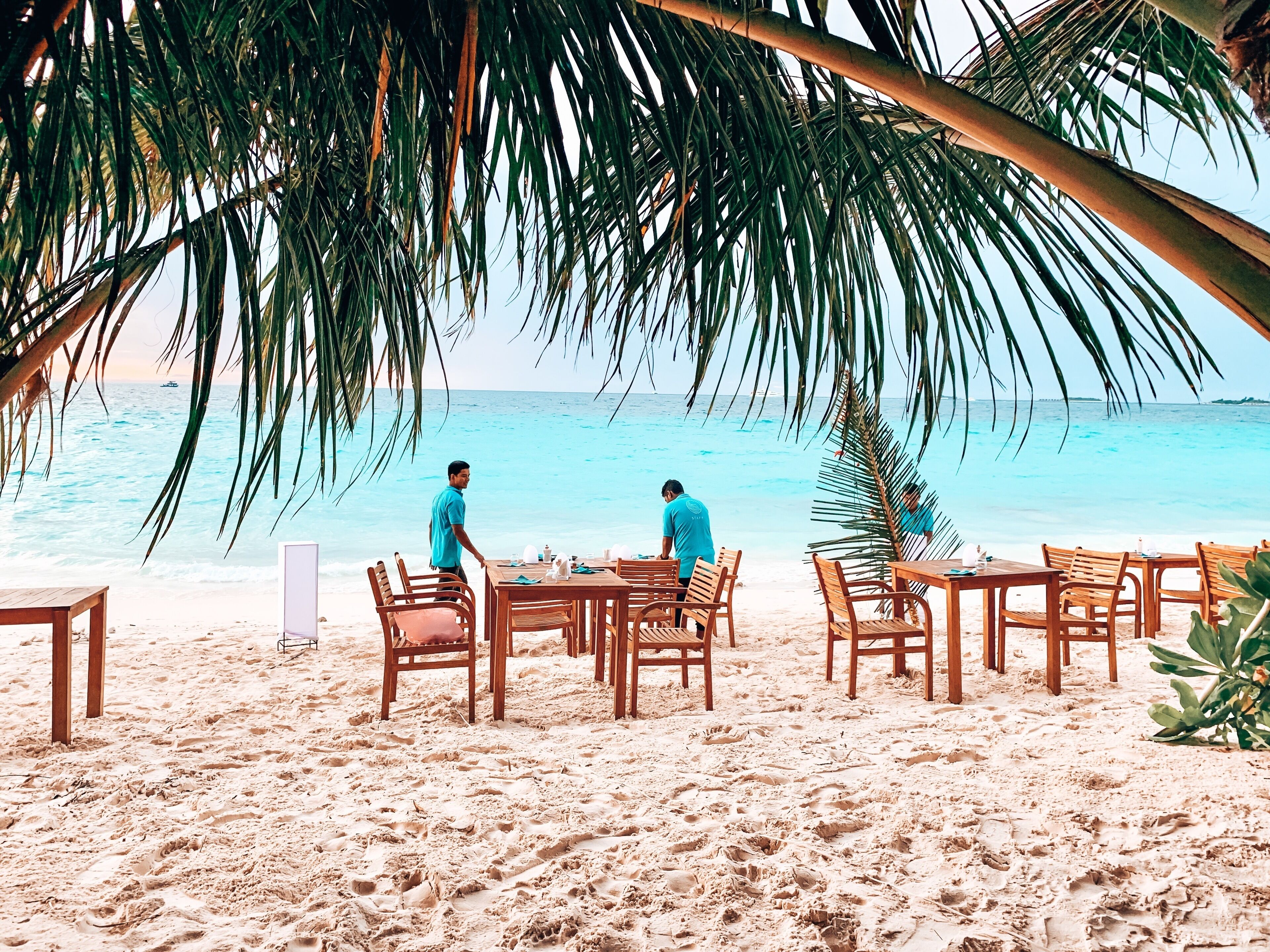 Un área de comedor frente a la playa, con mesas y sillas de madera, con vista al océano.