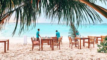 Un área de comedor frente a la playa, con mesas y sillas de madera, con vista al océano.