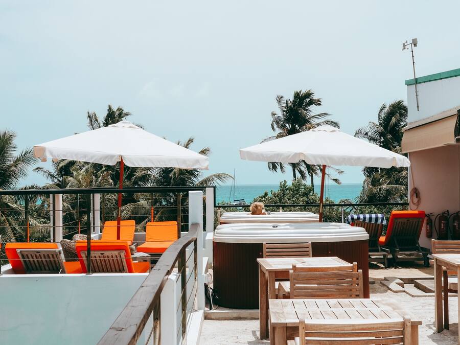 A rooftop terrace with wooden tables and orange lounge chairs, white umbrellas, and a hot tub.