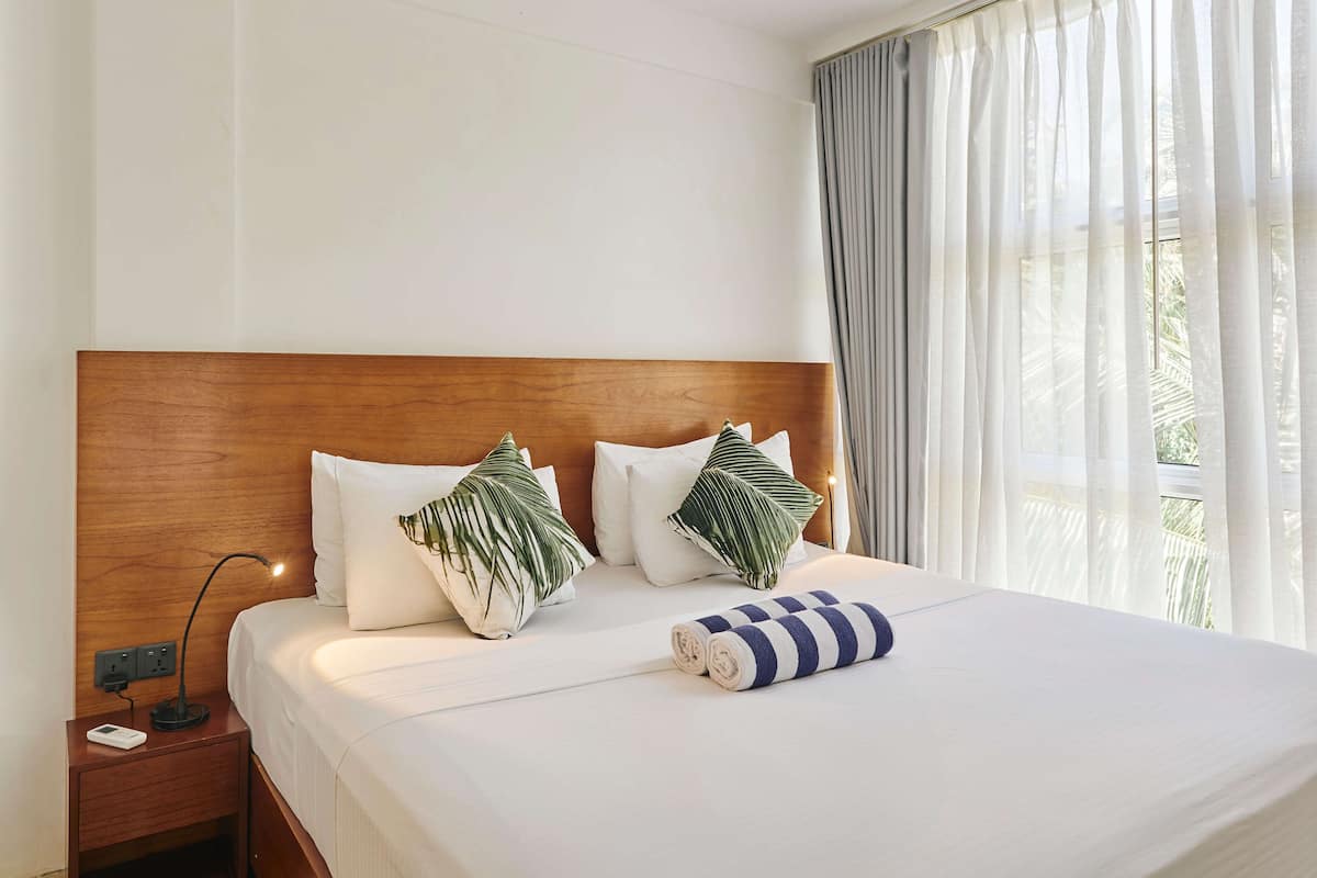 A hotel room with a wooden headboard, white bedding, two decorative pillows, a rolled towel, and a nightstand with a power outlet.