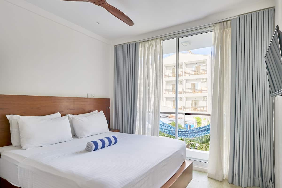 A hotel room with a bed, a ceiling fan, large windows with curtains, and a view of a building outside.