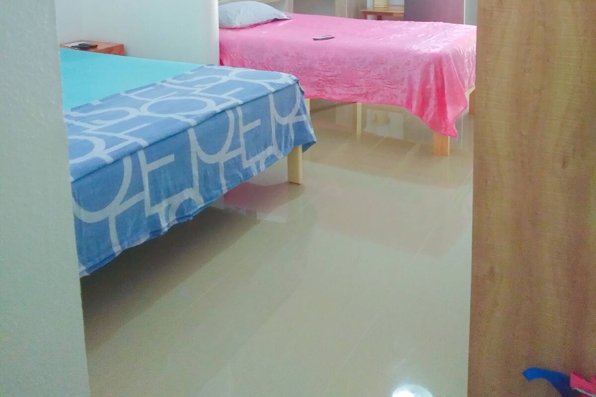 A small, well-lit room with two beds, one with a blue bedspread and the other with a pink bedspread. There is a small kitchenette area with a refrigerator and a sink.