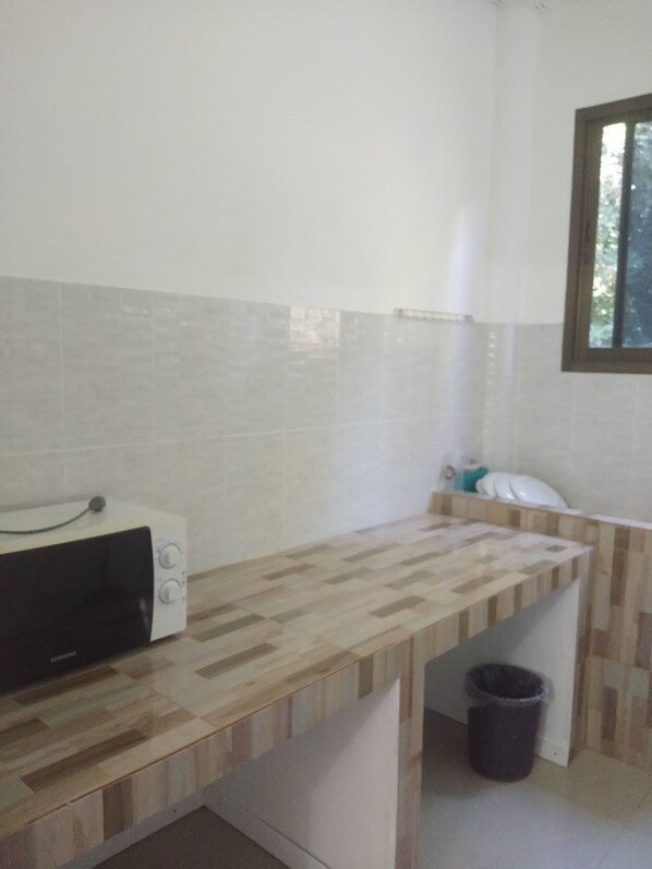 A kitchen with a microwave, a wooden countertop, a trash bin, and a window with a view of trees. - Chomlay Restaurant & Room for Rent (Rayong)