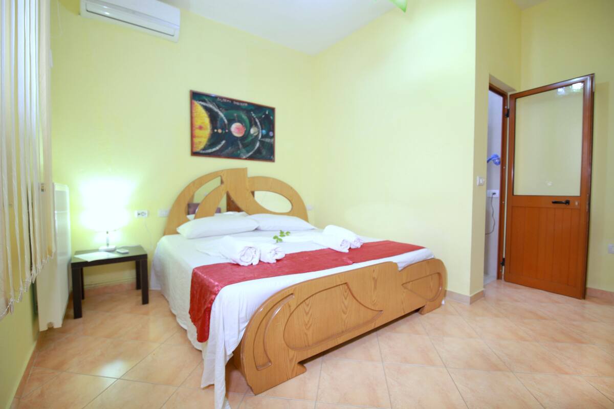 Un dormitorio con cama de madera, mesita de noche, aire acondicionado instalado en la pared, una puerta y un cuadro.