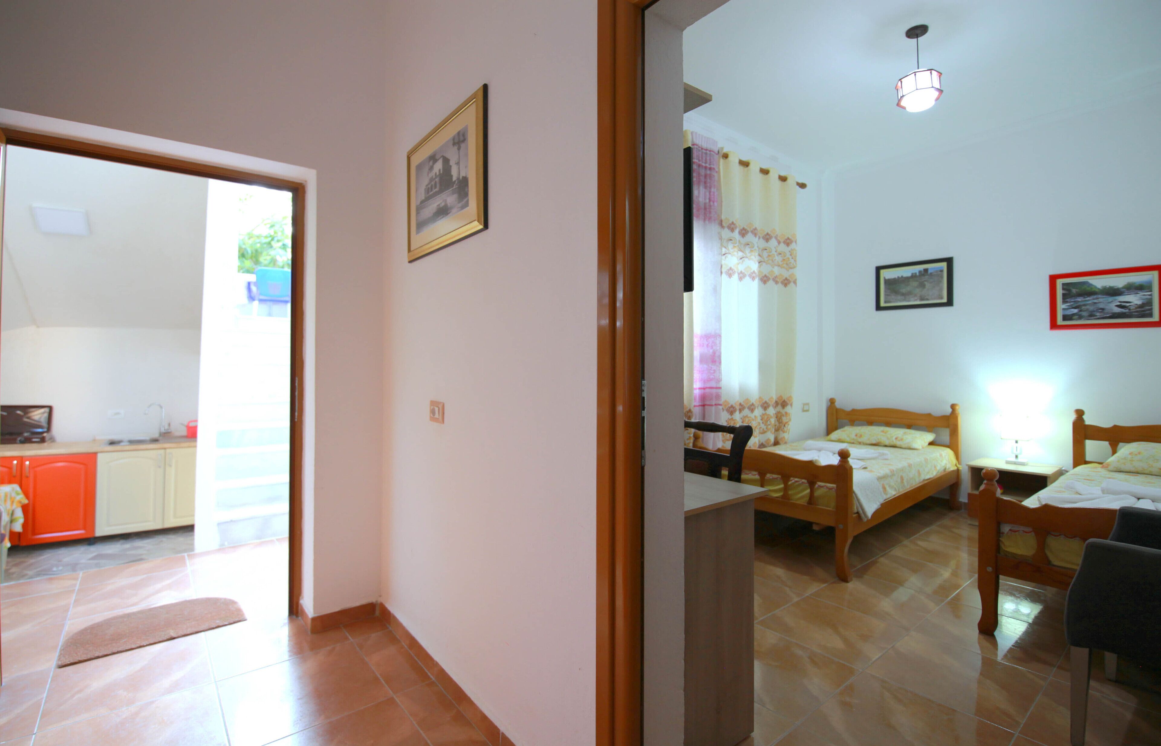 Una habitación con dos camas de madera, una mesita y una silla. Hay una ventana con cortinas, un cuadro en la pared y una puerta que da a otra habitación.