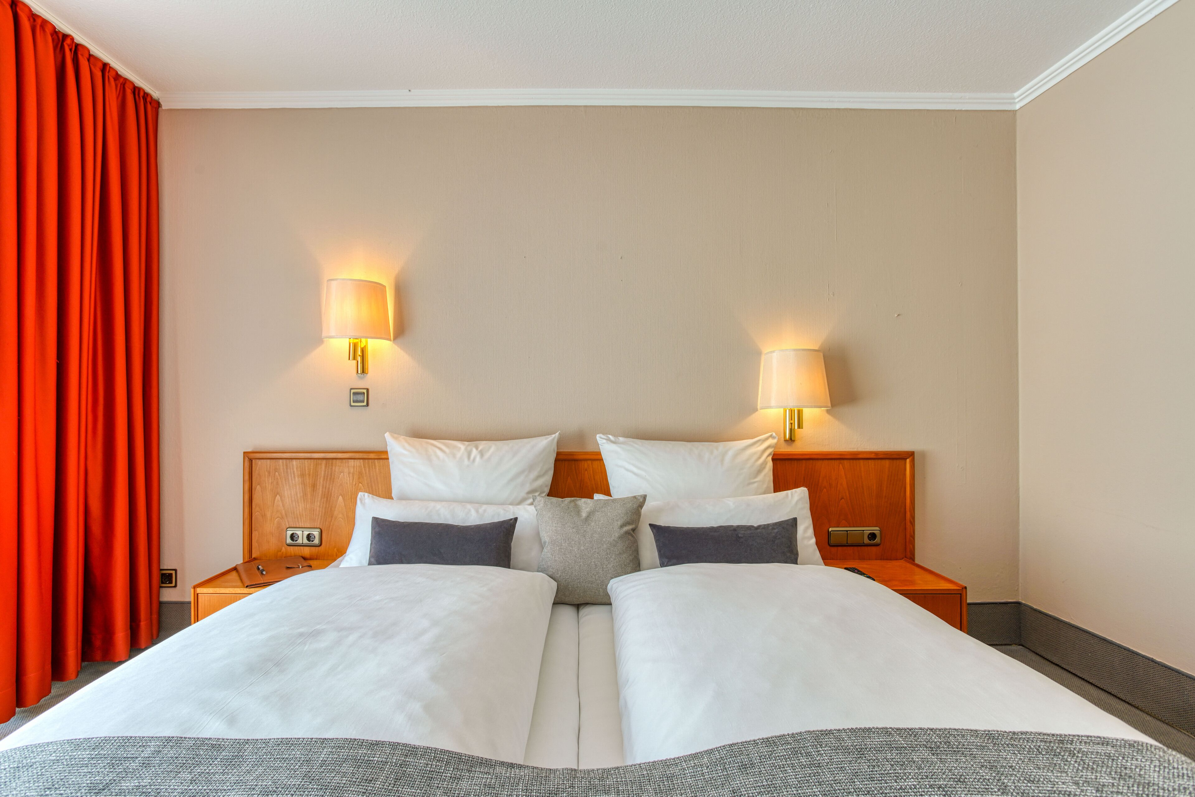 A double bed with white linens, two bedside tables, two wall-mounted lamps, and a red curtain.