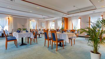 Un comedor de hotel con varias mesas y sillas, amplias ventanas con cortinas y una planta en maceta.