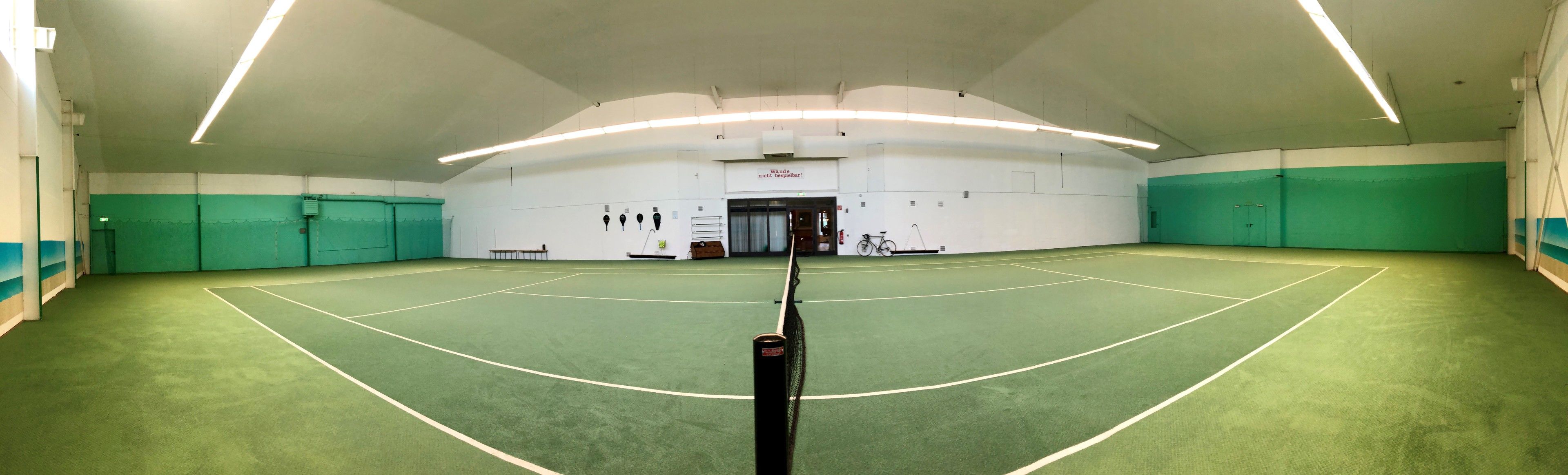 En indendørs tennisbane med et net i midten, omgivet af grønne vægge og hvide lofter.