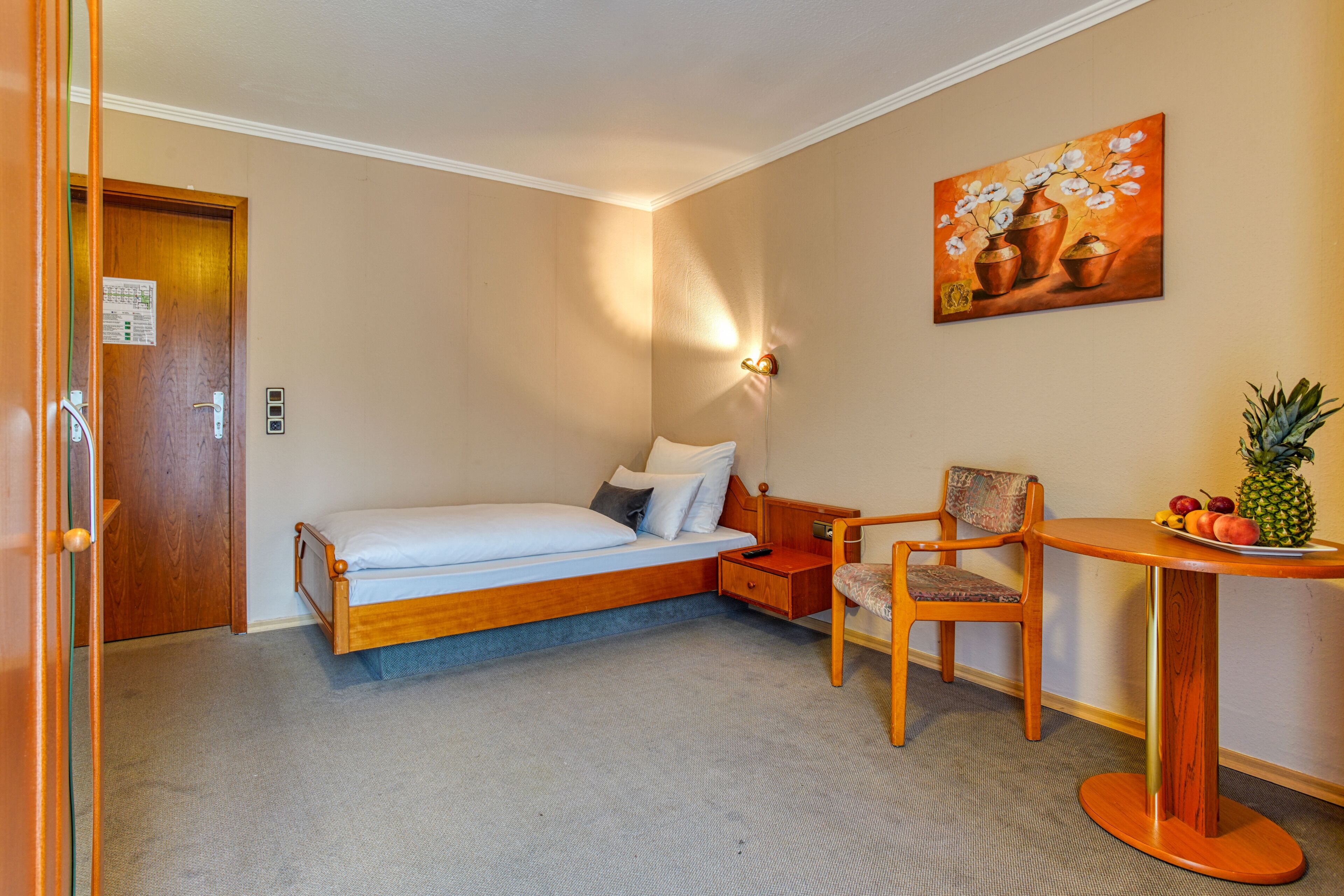 A hotel room with a bed, a chair, a small table with fruit, and a painting on the wall.