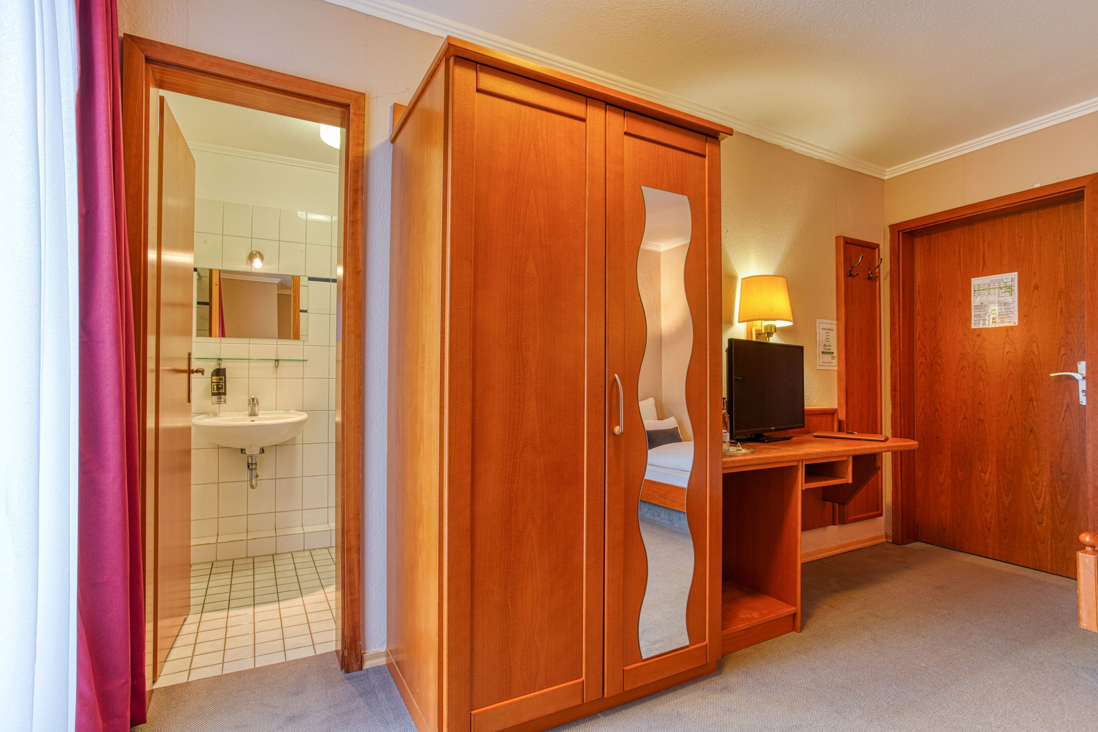 Een hotelkamer met een houten garderobe, een televisie, een bureau en een badkamer met een wastafel en spiegel.