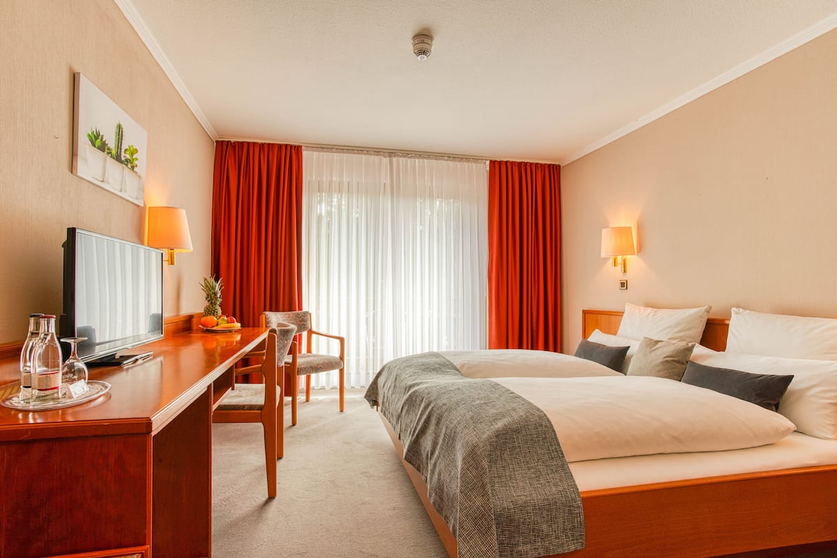 Ein Hotelzimmer mit einem Bett, einem Schreibtisch mit Fernseher, einem Sessel und einem Fenster mit Vorhängen.